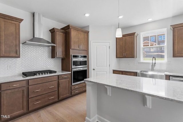 kitchen featuring wall chimney exhaust hood, stainless steel appliances, a sink, light wood-style floors, and a breakfast bar