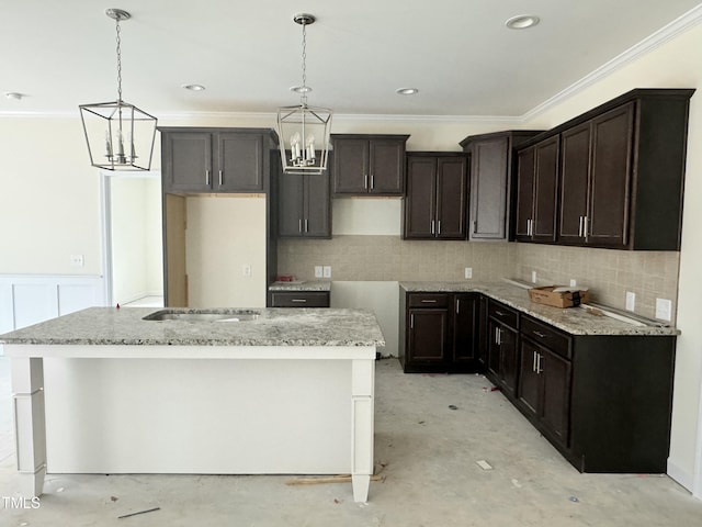 kitchen with light stone counters, ornamental molding, an island with sink, a notable chandelier, and pendant lighting