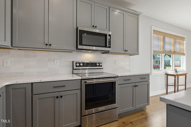 kitchen with tasteful backsplash, light hardwood / wood-style flooring, appliances with stainless steel finishes, gray cabinets, and light stone countertops