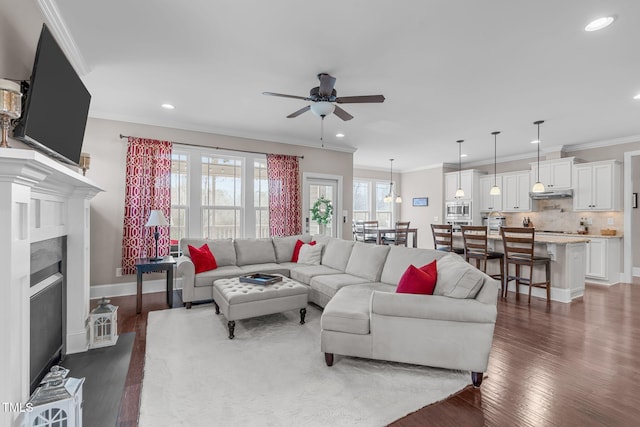 living room featuring ceiling fan, recessed lighting, dark wood-type flooring, a fireplace with flush hearth, and crown molding