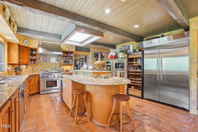 kitchen featuring beamed ceiling, appliances with stainless steel finishes, range hood, sink, and a breakfast bar