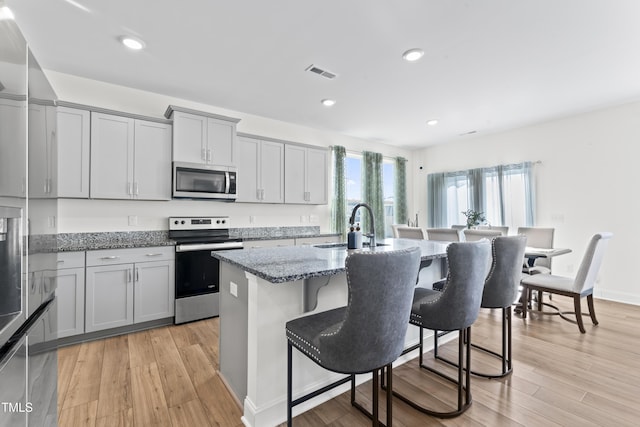 kitchen with sink, appliances with stainless steel finishes, gray cabinets, light stone countertops, and a kitchen island with sink