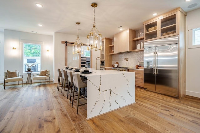 kitchen with a kitchen island with sink, light stone countertops, decorative light fixtures, stainless steel built in fridge, and light wood-type flooring