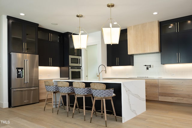 kitchen featuring appliances with stainless steel finishes, pendant lighting, decorative backsplash, a kitchen island with sink, and light hardwood / wood-style flooring