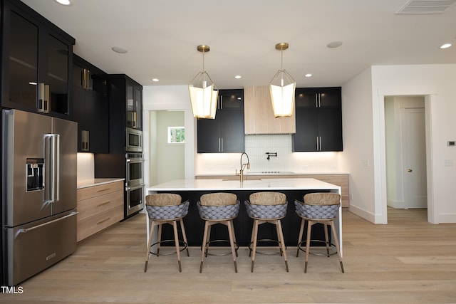 kitchen featuring appliances with stainless steel finishes, a kitchen island with sink, tasteful backsplash, decorative light fixtures, and light wood-type flooring