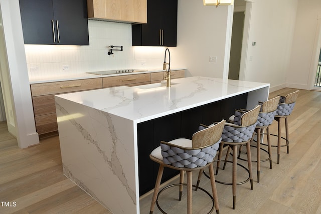 kitchen with light stone counters, black electric cooktop, a center island with sink, and light wood-type flooring