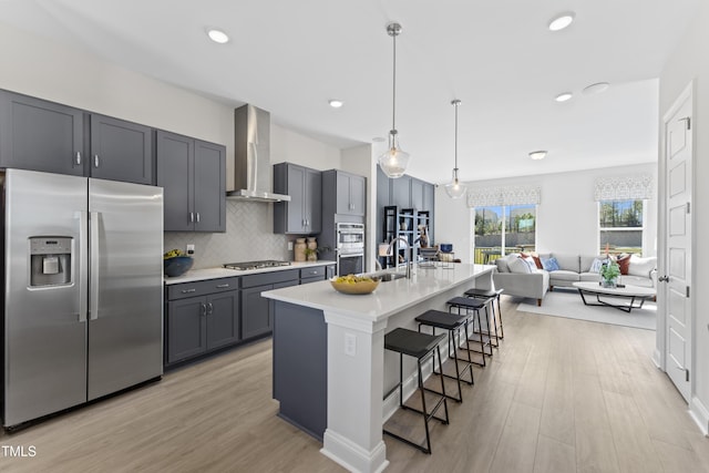 kitchen featuring a kitchen bar, gray cabinetry, stainless steel appliances, a kitchen island with sink, and wall chimney range hood