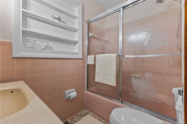 full bathroom with built in shelves, toilet, sink, bath / shower combo with glass door, and tile walls