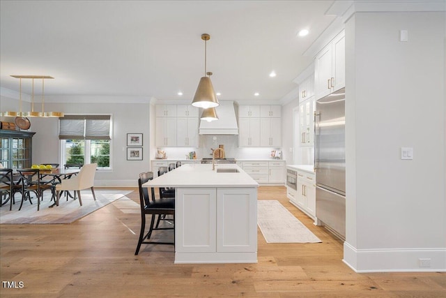 kitchen featuring pendant lighting, white cabinets, a kitchen island with sink, built in appliances, and wall chimney range hood