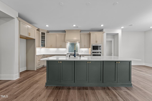 kitchen with premium range hood, light wood-type flooring, appliances with stainless steel finishes, a kitchen island with sink, and cream cabinetry