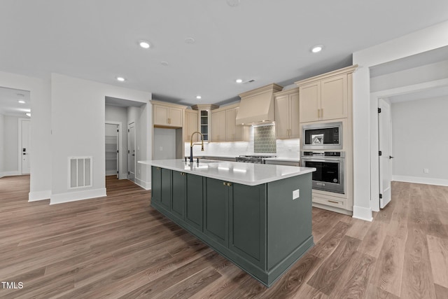 kitchen featuring appliances with stainless steel finishes, an island with sink, sink, light hardwood / wood-style floors, and custom range hood