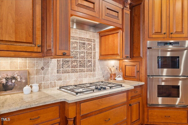 kitchen featuring brown cabinetry, decorative backsplash, light stone counters, stainless steel appliances, and wall chimney range hood