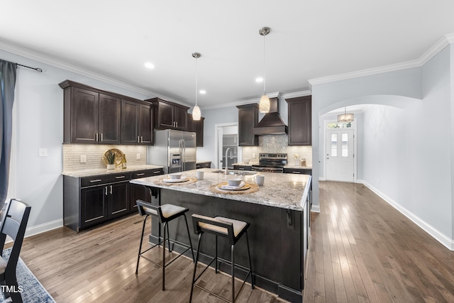 kitchen with arched walkways, stainless steel appliances, a sink, wall chimney exhaust hood, and an island with sink