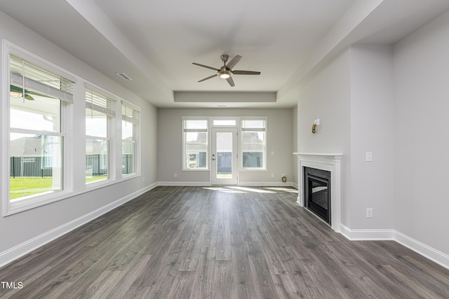 unfurnished living room with a raised ceiling, visible vents, a glass covered fireplace, dark wood finished floors, and a ceiling fan