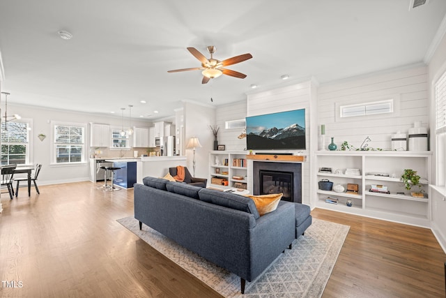 living room featuring light wood finished floors, baseboards, a glass covered fireplace, ceiling fan, and crown molding