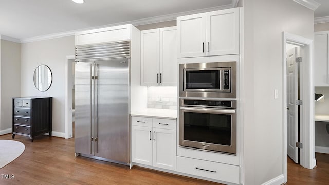 kitchen with decorative backsplash, ornamental molding, white cabinets, wood finished floors, and built in appliances