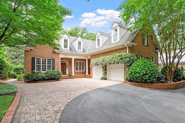 cape cod home with driveway, brick siding, a garage, covered porch, and a shingled roof