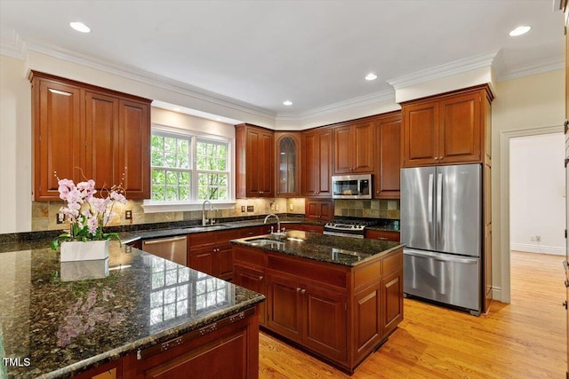 kitchen featuring stainless steel appliances, a sink, a center island with sink, light wood-style flooring, and dark stone countertops