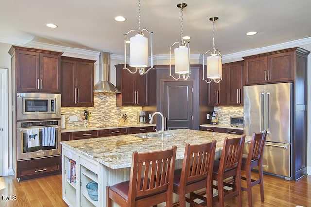 kitchen with appliances with stainless steel finishes, decorative light fixtures, an island with sink, sink, and wall chimney range hood