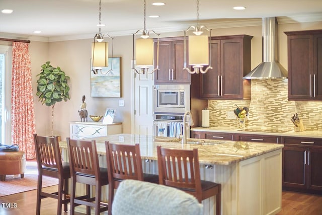 kitchen with sink, light stone counters, a center island with sink, stainless steel appliances, and wall chimney range hood