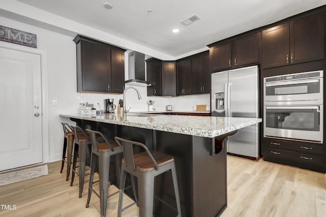 kitchen with dark brown cabinetry, light stone countertops, stainless steel appliances, and wall chimney exhaust hood