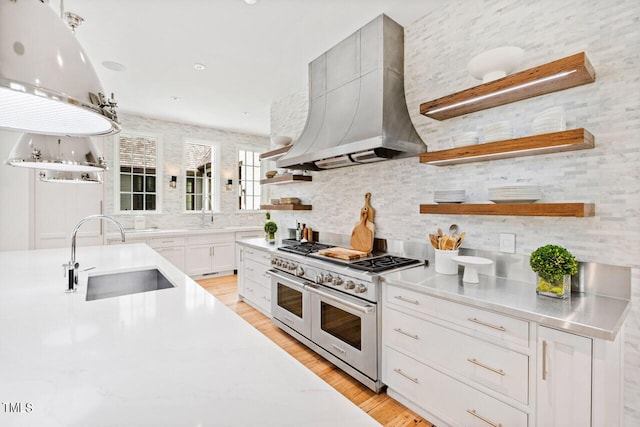 kitchen with sink, wall chimney range hood, light hardwood / wood-style floors, white cabinets, and range with two ovens