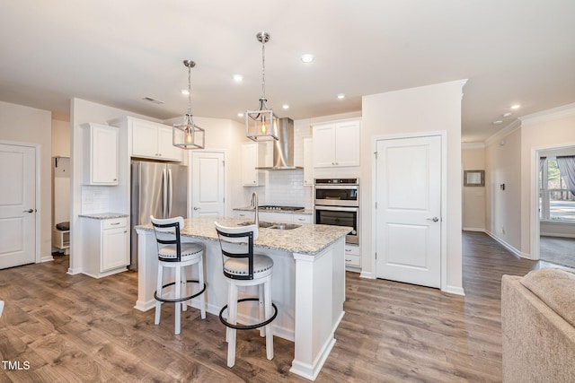 kitchen with tasteful backsplash, white cabinets, stainless steel appliances, a center island with sink, and wall chimney range hood