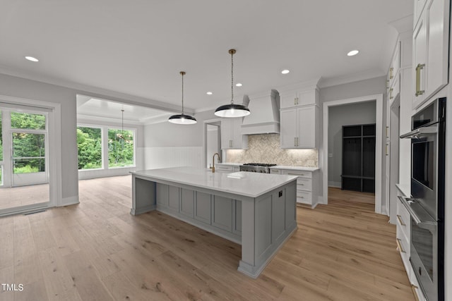 kitchen featuring premium range hood, ornamental molding, an island with sink, white cabinets, and decorative light fixtures