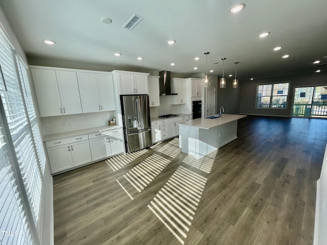 kitchen with pendant lighting, an island with sink, white cabinets, stainless steel fridge, and wall chimney range hood