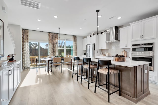 kitchen with appliances with stainless steel finishes, pendant lighting, wall chimney range hood, a kitchen island with sink, and white cabinets