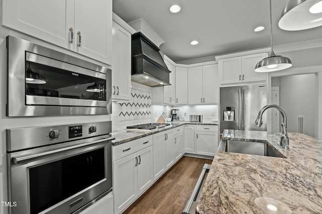 kitchen with premium range hood, appliances with stainless steel finishes, white cabinetry, sink, and hanging light fixtures