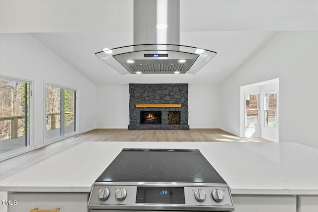 kitchen with stainless steel electric range, vaulted ceiling, a stone fireplace, island exhaust hood, and light hardwood / wood-style flooring