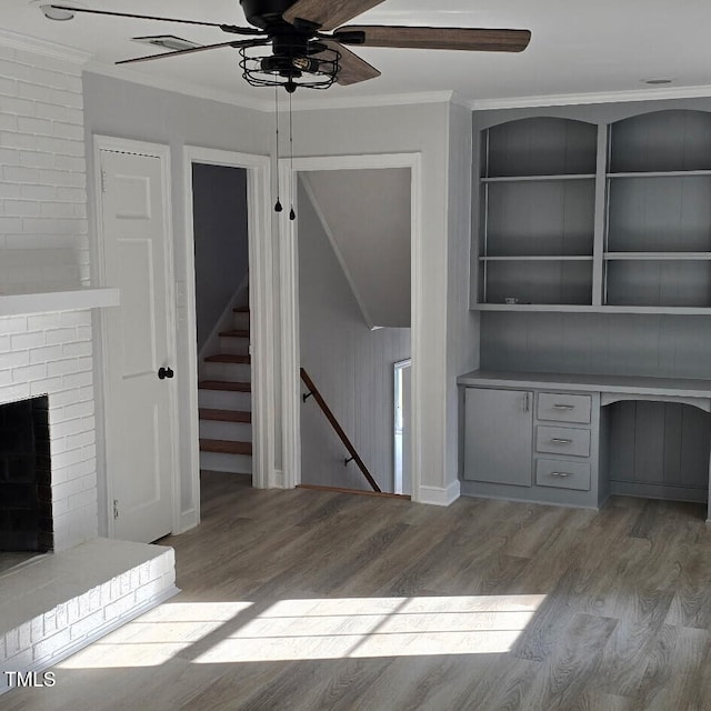 unfurnished office featuring built in desk, a brick fireplace, ornamental molding, hardwood / wood-style flooring, and ceiling fan