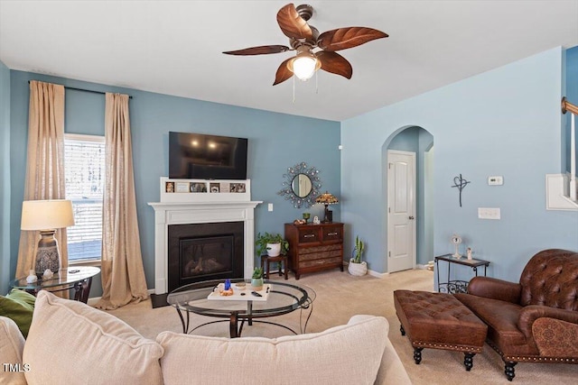 living room featuring arched walkways, light colored carpet, a ceiling fan, a glass covered fireplace, and baseboards