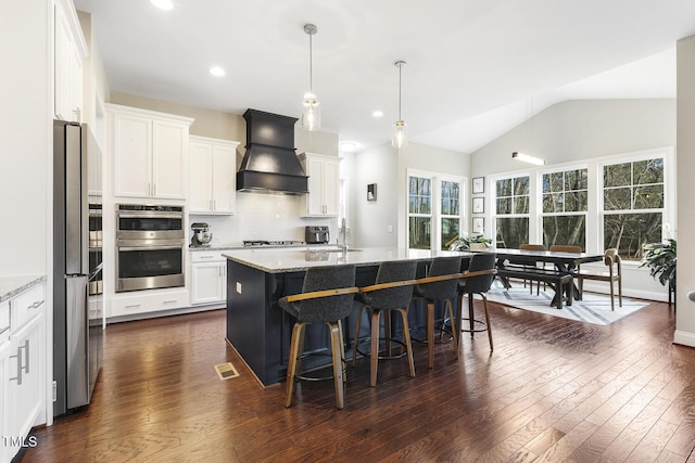 kitchen featuring a center island with sink, light stone counters, custom range hood, white cabinets, and a kitchen bar