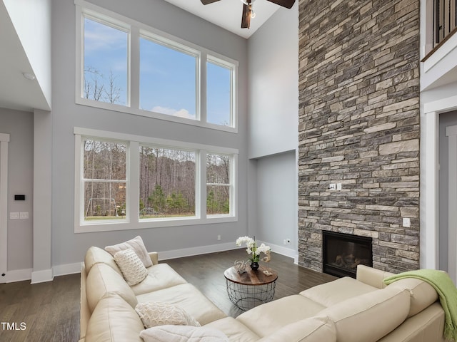 living area featuring dark wood-style floors, baseboards, ceiling fan, a stone fireplace, and a towering ceiling