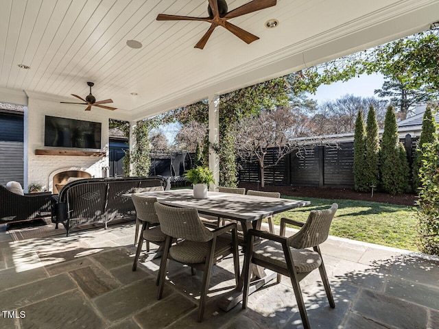 view of patio with ceiling fan, a lit fireplace, outdoor dining space, and a fenced backyard