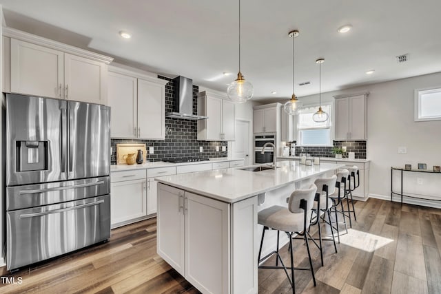 kitchen featuring decorative light fixtures, an island with sink, sink, stainless steel appliances, and wall chimney range hood