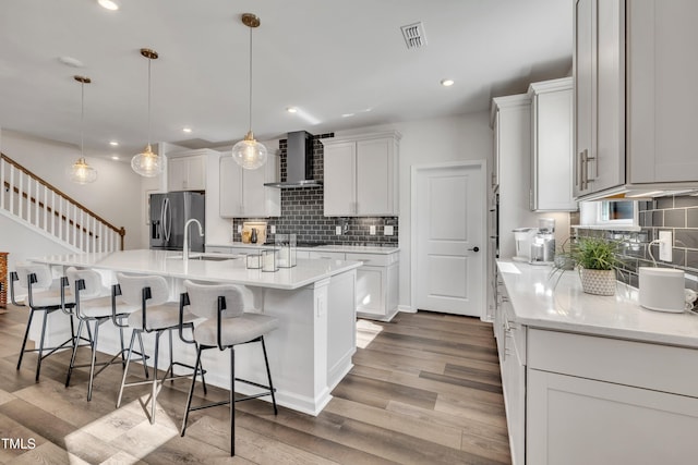 kitchen featuring an island with sink, white cabinetry, hanging light fixtures, stainless steel refrigerator with ice dispenser, and wall chimney exhaust hood