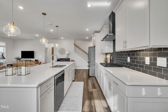 kitchen with sink, wall chimney range hood, appliances with stainless steel finishes, hanging light fixtures, and a large island with sink