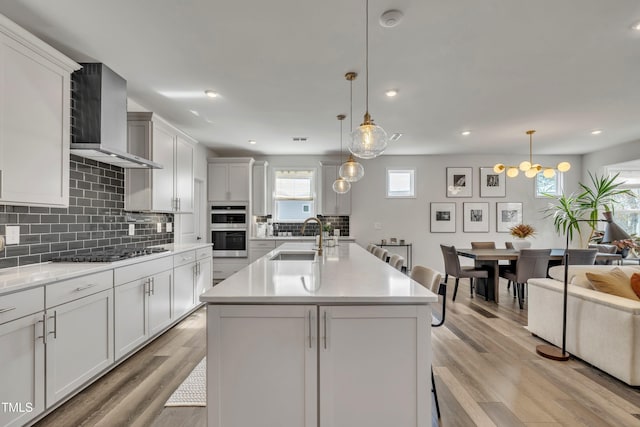 kitchen featuring sink, appliances with stainless steel finishes, a kitchen island with sink, hanging light fixtures, and wall chimney exhaust hood