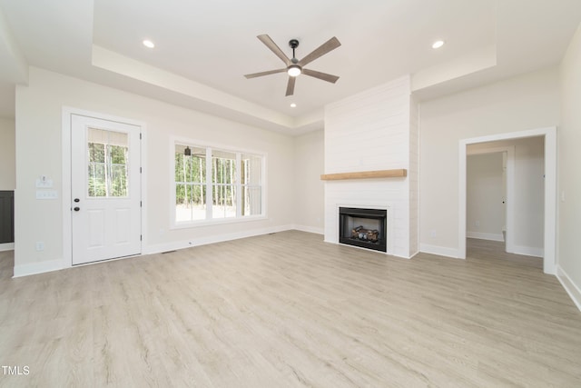 unfurnished living room with ceiling fan, a large fireplace, a raised ceiling, and light wood-type flooring