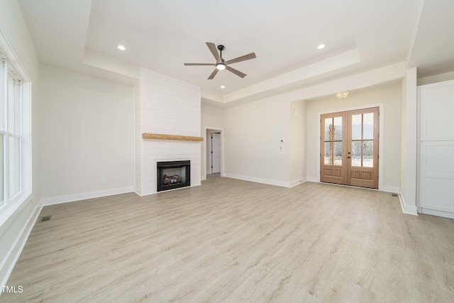 unfurnished living room featuring french doors, light hardwood / wood-style flooring, a large fireplace, a raised ceiling, and ceiling fan