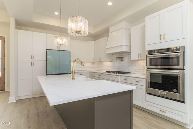 kitchen with light stone counters, a center island with sink, a tray ceiling, stainless steel appliances, and white cabinets