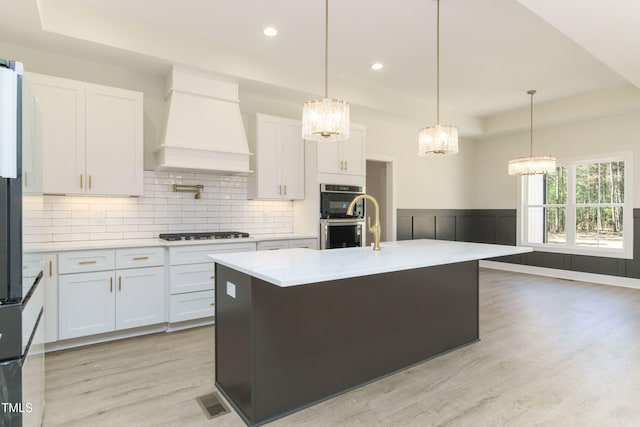 kitchen featuring premium range hood, a raised ceiling, a kitchen island with sink, and white cabinets