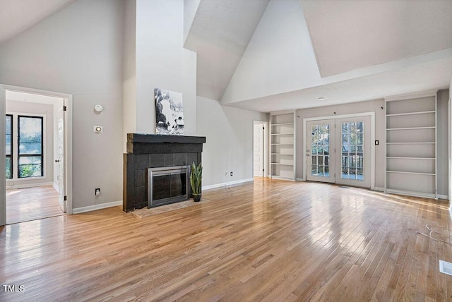 unfurnished living room with french doors, high vaulted ceiling, built in features, a fireplace, and light hardwood / wood-style floors