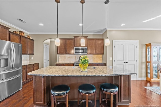 kitchen with appliances with stainless steel finishes, an island with sink, hanging light fixtures, light stone counters, and dark wood-type flooring