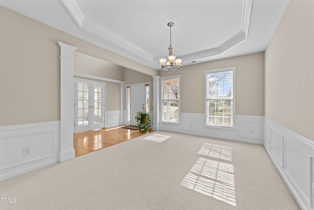 carpeted empty room with french doors, ornate columns, crown molding, a tray ceiling, and a notable chandelier