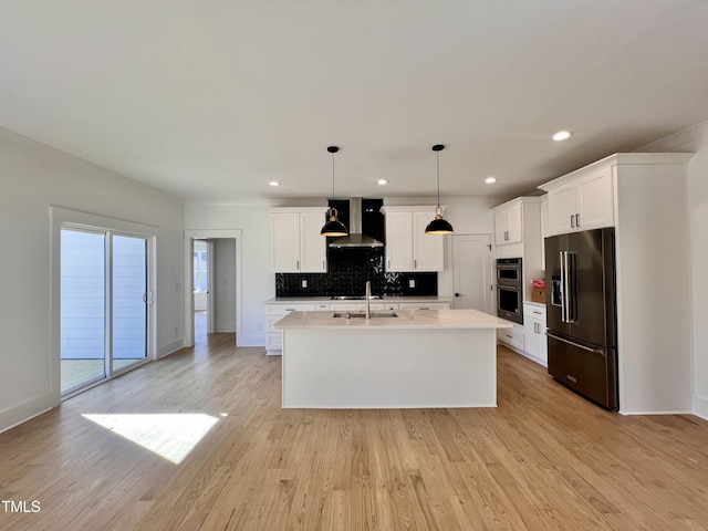 kitchen featuring hanging light fixtures, high end black fridge, wall chimney exhaust hood, sink, and a kitchen island with sink