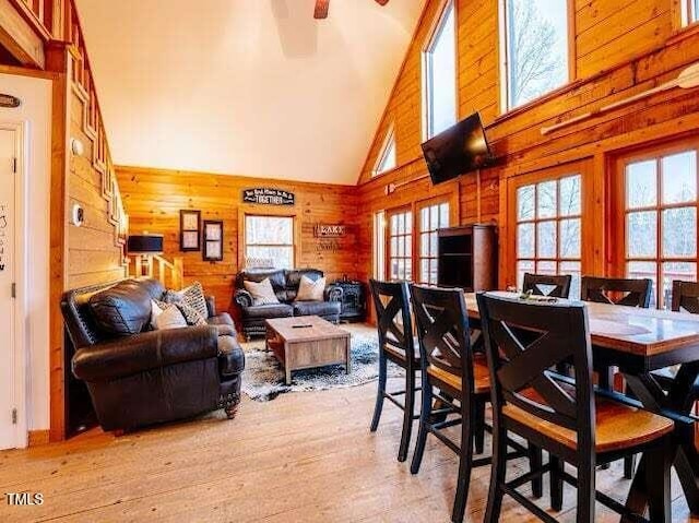 dining space featuring ceiling fan, high vaulted ceiling, light wood-type flooring, and wooden walls